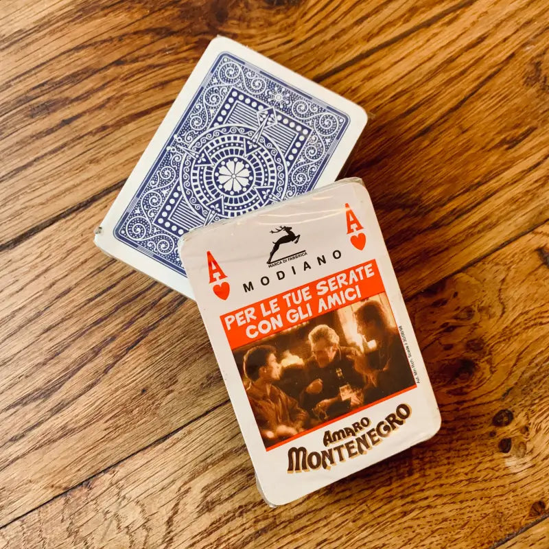 Carte da gioco con dorso a fantasia blu e cartoncino promozionale del liquore ’Amaro Montenegro’.
