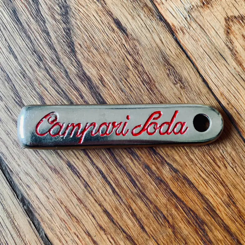 Portachiavi o targhetta in metallo con scritta ’Campari Soda’ in caratteri rossi.