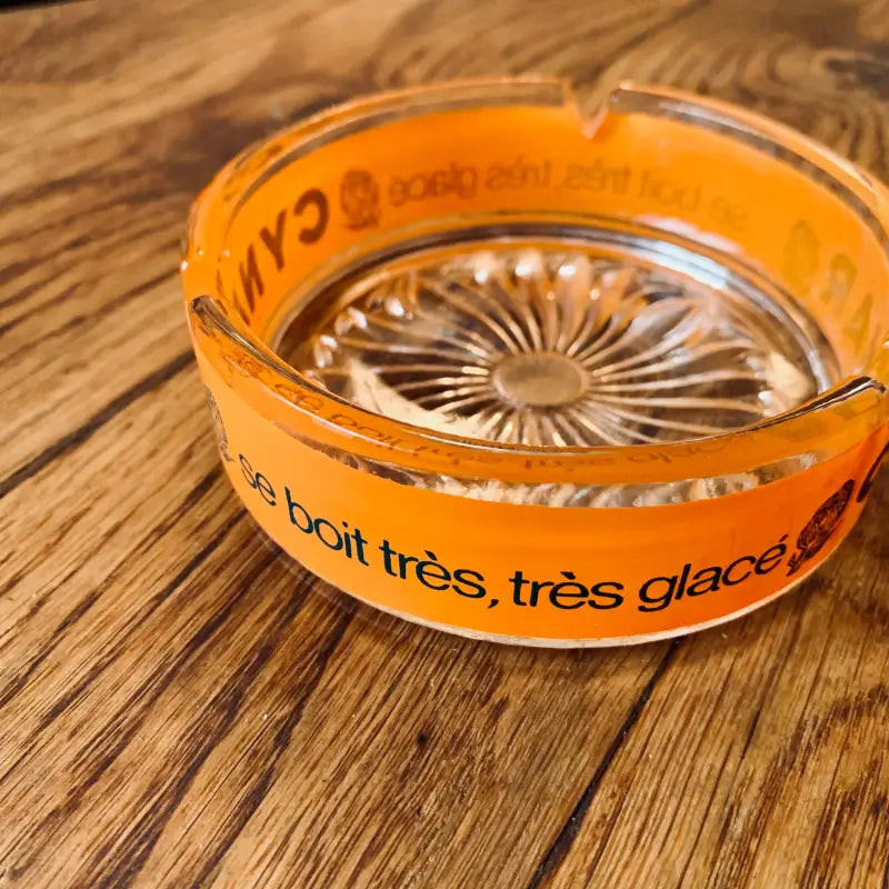 Posacenere in vetro arancione con testo in francese e motivo decorativo sul fondo.