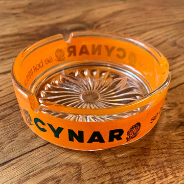 Posacenere in vetro arancione con marchio ’CYNAR’ e fondo decorativo in vetro tagliato.